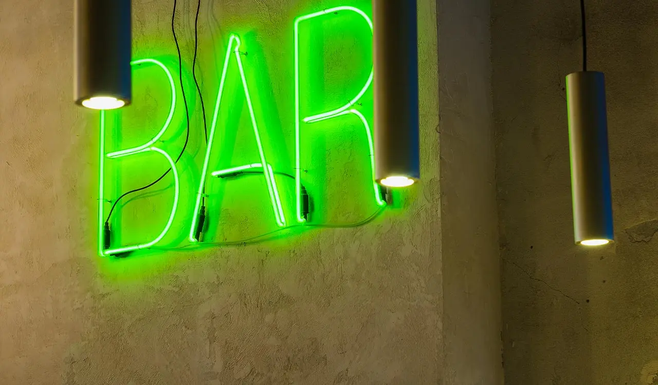 Grüner Neonschriftzug „BAR“ auf rauer Wand zwischen Pendelleuchten