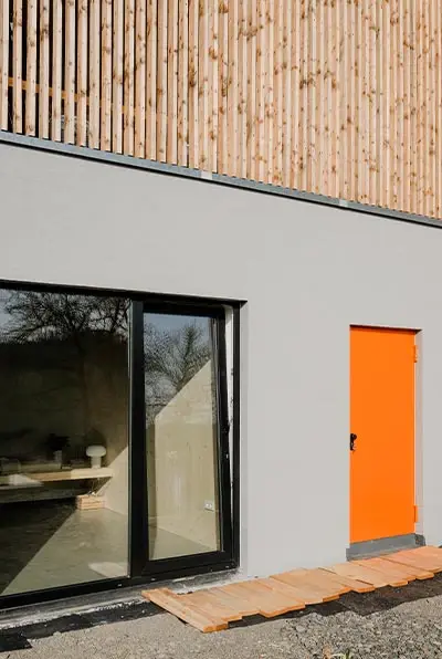 Gartenseite mit großen Fenstern und orangefarbener Tür unter der Holzlamellenfassade